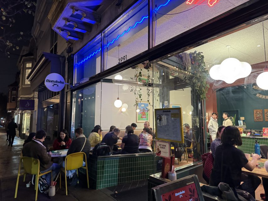 A group of people sit at tables inside a brightly lit restaurant with large windows and a neon sign.