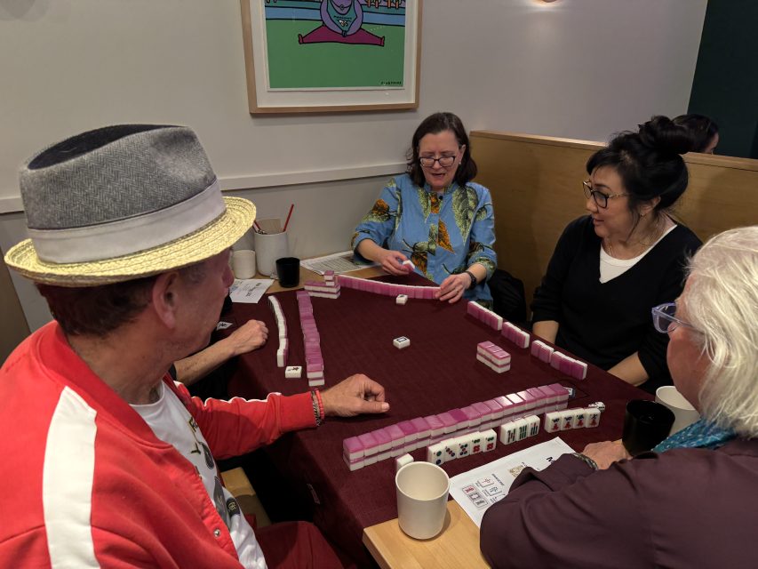 Four people play mahjong around a table. The woman in blue is smiling while holding a tile. Cups and tiles are on the table.