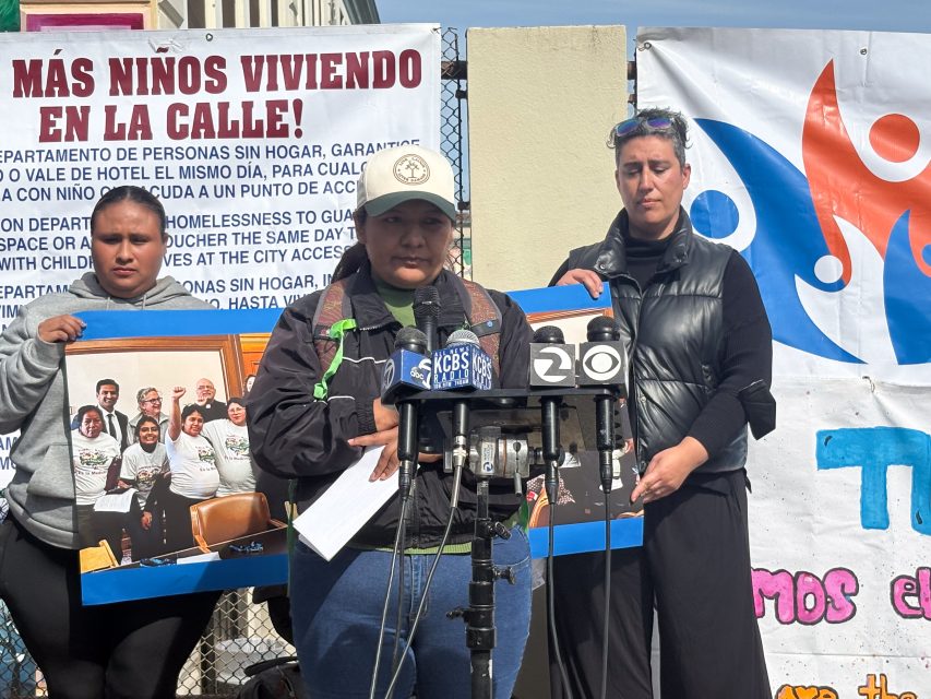 A group of people stand in front of a banner and microphones at a press event. One person holds a photo of a group, and another speaks into the microphones.