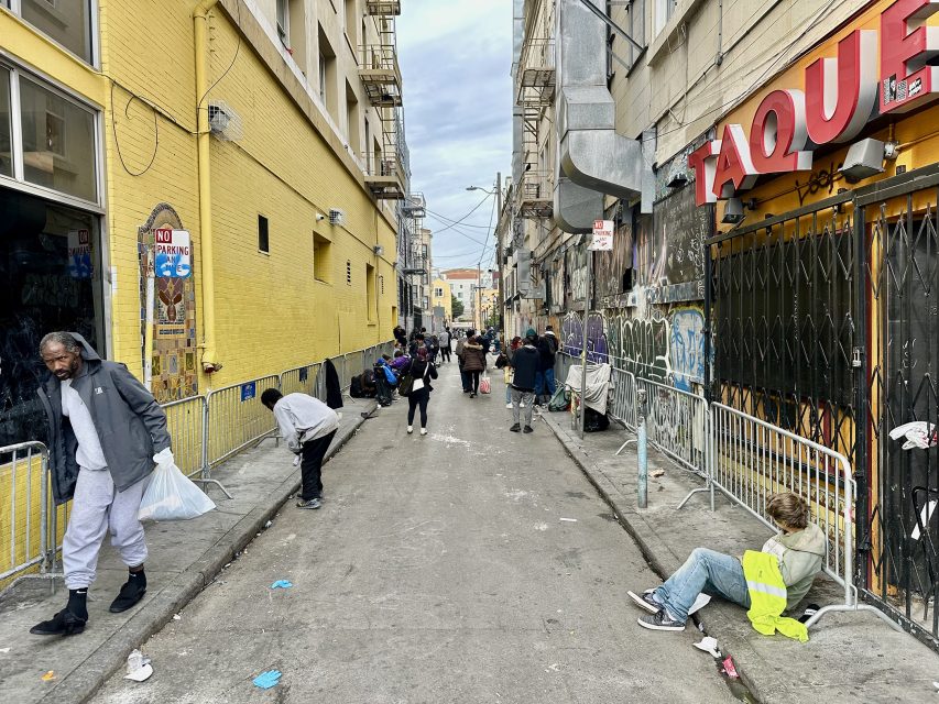 Narrow alley with people standing or sitting, some with belongings, against graffiti-covered walls and metal barriers. A yellow building and a taco shop are on either side. Overcast sky above.