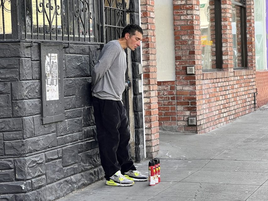 A man leans against the black and brick wall at 16th St. Plaza, gazing downward, with spray paint cans scattered on the ground before him.