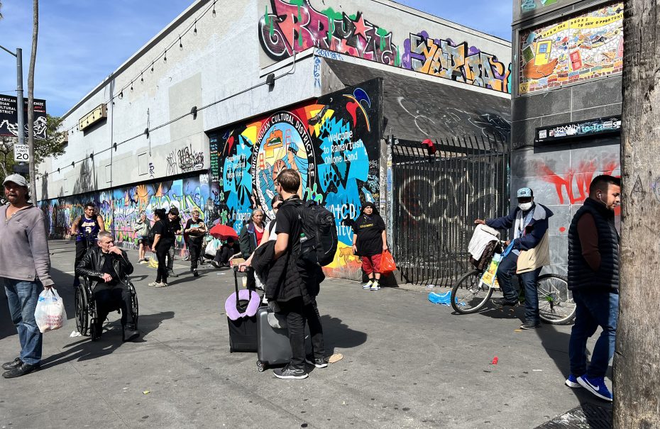 A group of people stands on the graffiti-covered sidewalk at 16th St. Plaza. Some carry bags or luggage, and nearby, a bicycle leans against a wall. A person in a wheelchair accompanies them, with a tall wooden pole marking the scene on the right.