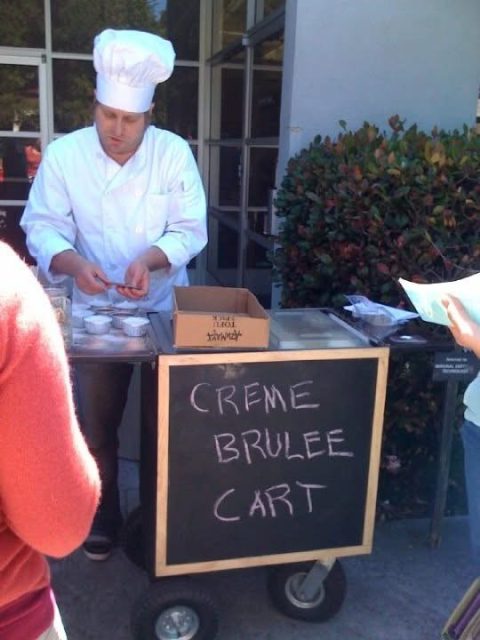 Curtis Kimball, the chef renowned for elevating street cuisine, elegantly serves crème brûlée from a cart outdoors, dressed in his signature white uniform and hat.