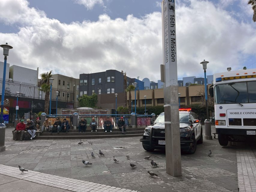 Urban plaza scene with a police car, pigeons on the ground, and people sitting on benches near a sign labeled "16th St Mission." A mobile command center vehicle is parked nearby.