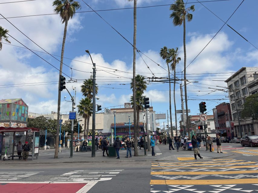 A busy street intersection with palm trees and people walking. Buildings line the background, and overhead power lines crisscross the sky.