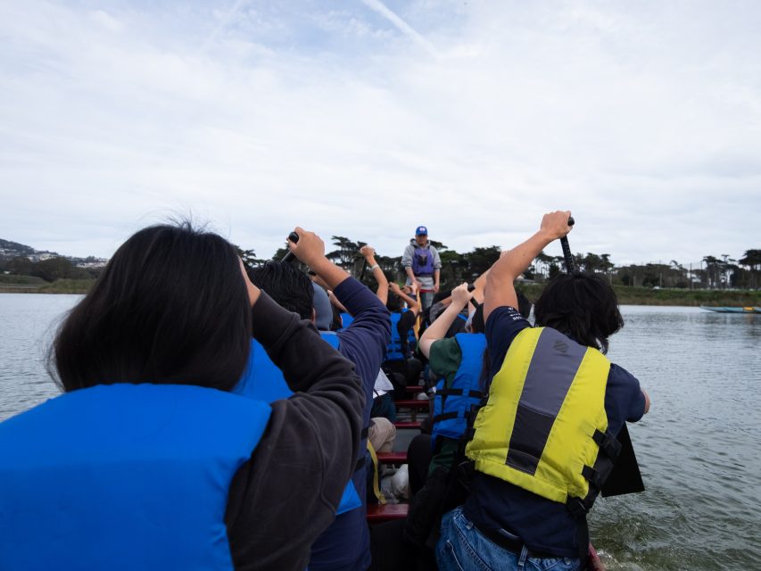 People rowing in a dragon boat on a lake, wearing blue and yellow life jackets, with trees visible in the background.