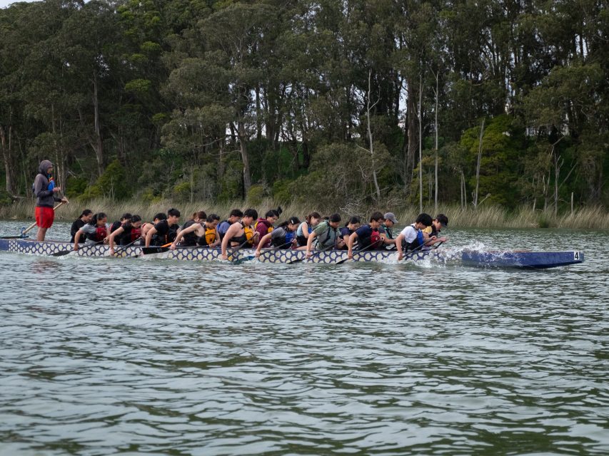A group of people paddling in a dragon boat on a calm lake, overseen by a person standing at the back. Trees line the shore in the background.