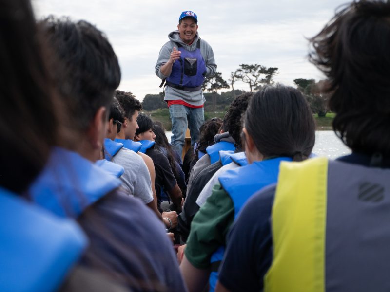 A person stands in a boat, speaking to a group of seated people wearing blue life jackets on a calm waterway. Trees and cloudy sky can be seen in the background.