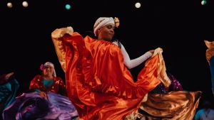 Dancer in vibrant costume performs on stage, holding orange and gold fabric, with colorful-clad individuals blurred in the background.