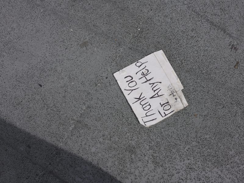 A piece of white paper with handwritten text "Thank you for any help" lies on a gray, textured ground.