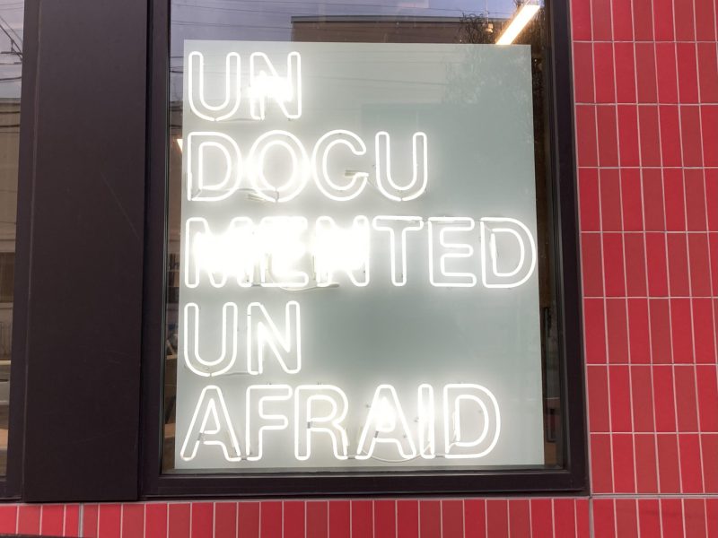 Neon sign in a window reads "UNDOCUMENTED UNAFRAID" against a red tiled wall.