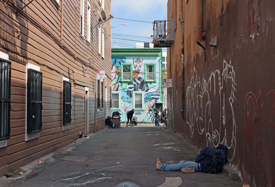 Narrow alleyway with graffiti on walls, leading to a colorful mural on a building. A person is lying on the ground in the foreground, with others further down the alley.