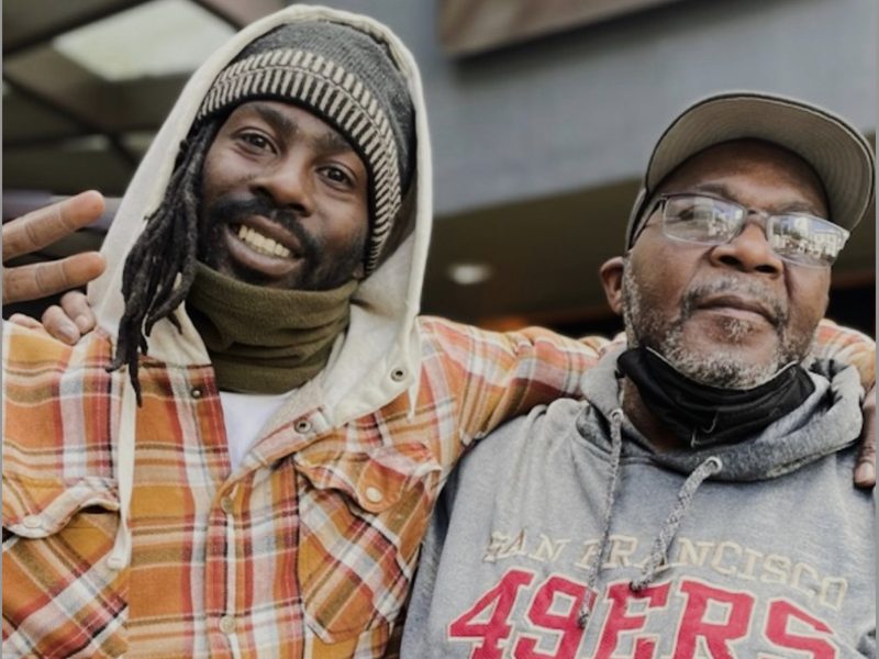Two men pose together outdoors; one wears a plaid shirt and knit cap, the other a 49ers hoodie and cap. Both smile at the camera.