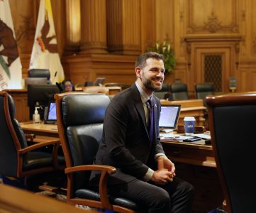 A person in a suit sits and smiles in a wood-paneled courtroom or government chamber with flags, a laptop, and a blue coffee cup.