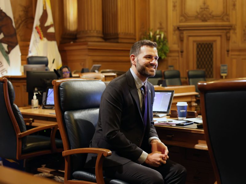 A person in a suit sits and smiles in a wood-paneled courtroom or government chamber with flags, a laptop, and a blue coffee cup.