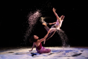Two dancers perform on a dark stage with scattered powder, one leaping in the air while the other extends a hand from the ground.