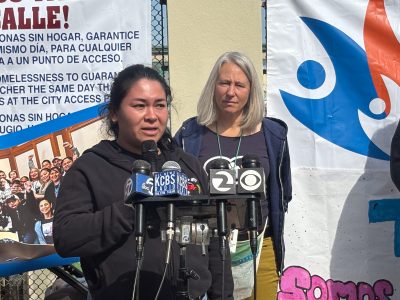 Two women stand at a podium with multiple microphones. One woman speaks while the other looks on. Behind them is a banner with text in English and Spanish and a blue and orange design.