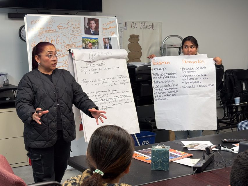 Two women present research findings in a meeting room, using flip charts with handwritten notes. A board with photos and diagrams is in the background.