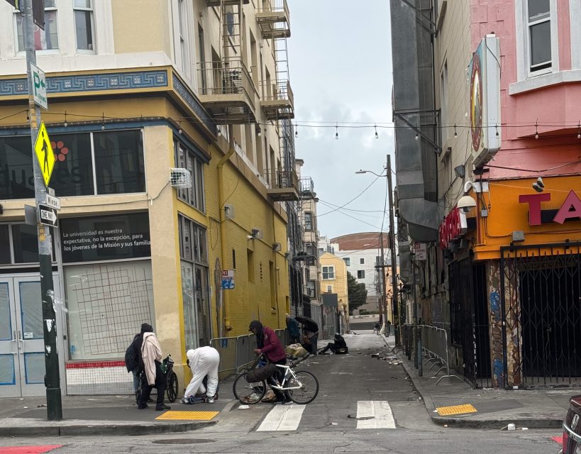 A narrow city street with three people near a crosswalk, one on a bicycle. Buildings line both sides, and a yellow storefront and taco restaurant are visible. Some debris on the ground.
