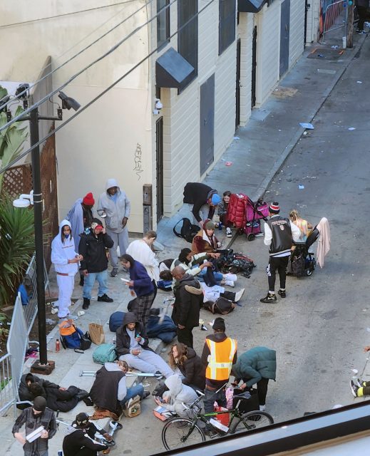 In a bustling alley, a group gathers beside a building, some perched on bicycles while others rest on the ground. A person in a reflective vest stands out near the bottom right, adding to the vibrant scene woven into this everyday tapestry.