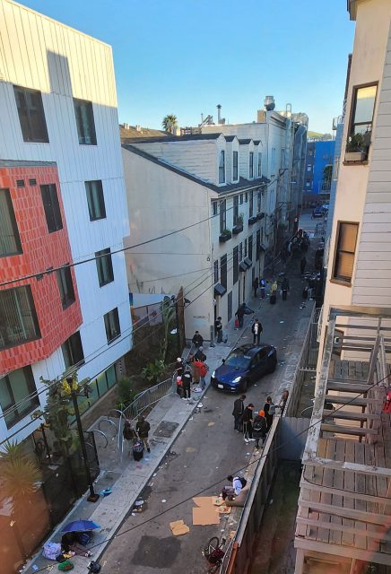 A narrow urban alley buzzes with people, scattered trash, and parked vehicles. The alleys are lined with buildings showcasing diverse architectural styles, basking under a clear blue sky.
