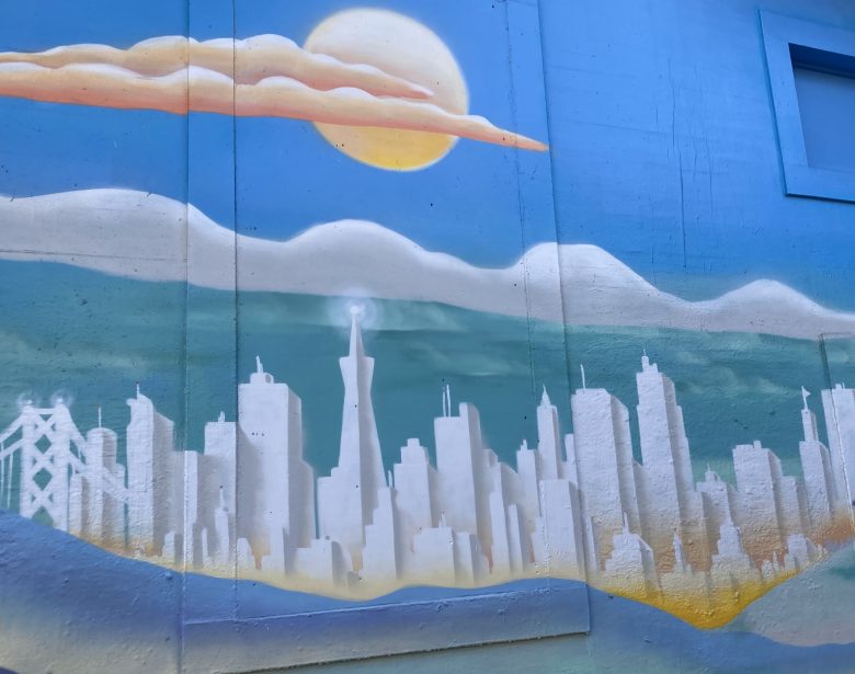 Mural of a white city skyline with a bridge, hills, and a large sun against a blue sky with clouds, painted on a wall.