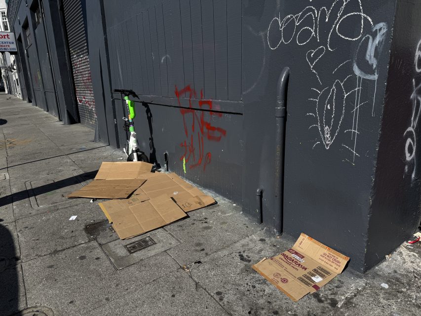 Several flattened cardboard boxes lie on a city sidewalk next to a dark gray wall with graffiti, a pipe, and a partially visible electric scooter.
