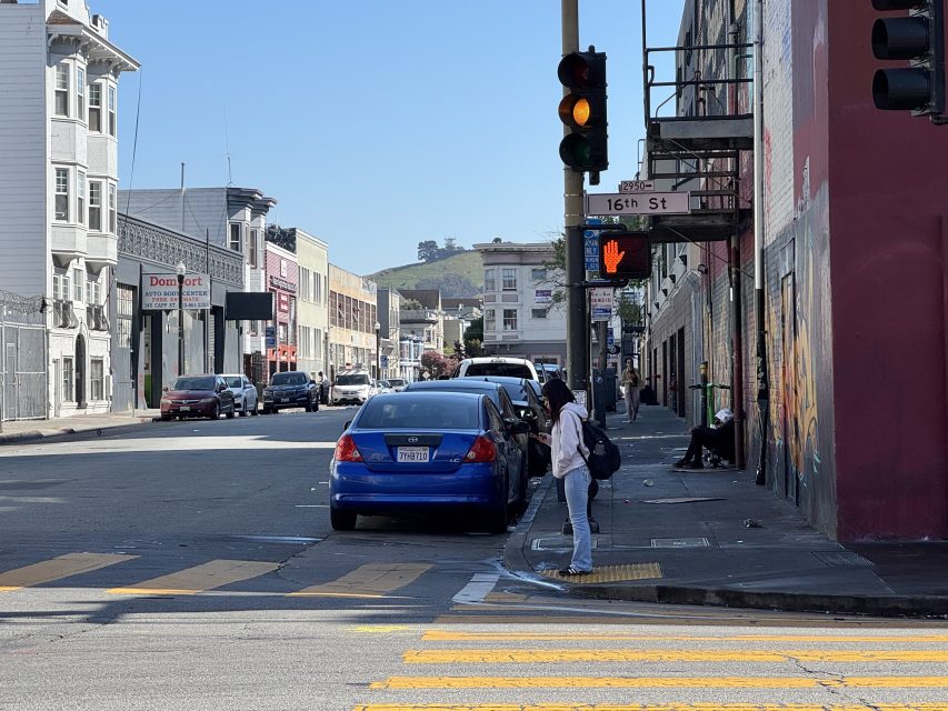 A woman stands at the corner of 16th Street, waiting to cross while the pedestrian signal shows a red hand. Cars are parked along the street on a sunny day.