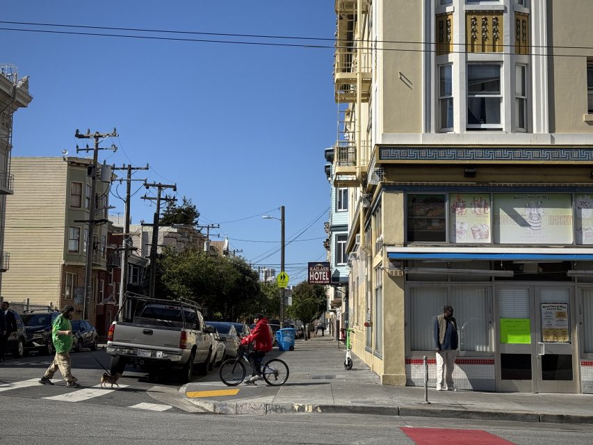 A person rides a bicycle past a hotel on a sunny street corner as others walk nearby; a pickup truck is parked at the curb.