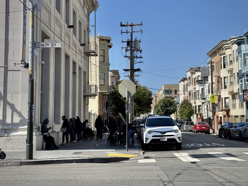 People gather on a street corner near a white building at the intersection of 16th Street; cars are parked along the road under a clear sky.