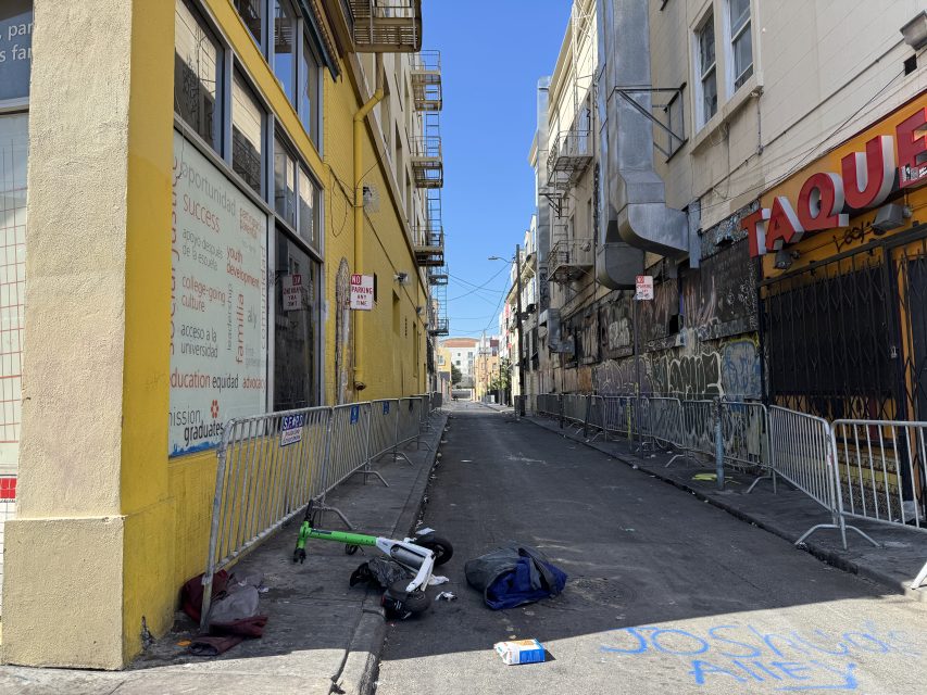 A narrow urban alleyway lined with metal barricades, graffiti-covered walls, scattered trash, and a discarded bicycle missing its front wheel.