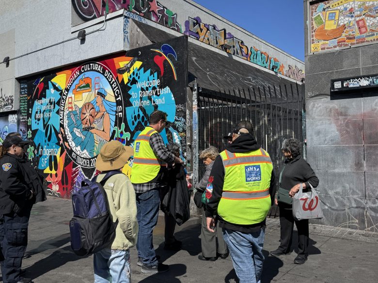 A group of people, some wearing yellow safety vests, gather near a colorful mural and graffiti-covered building in an urban area.