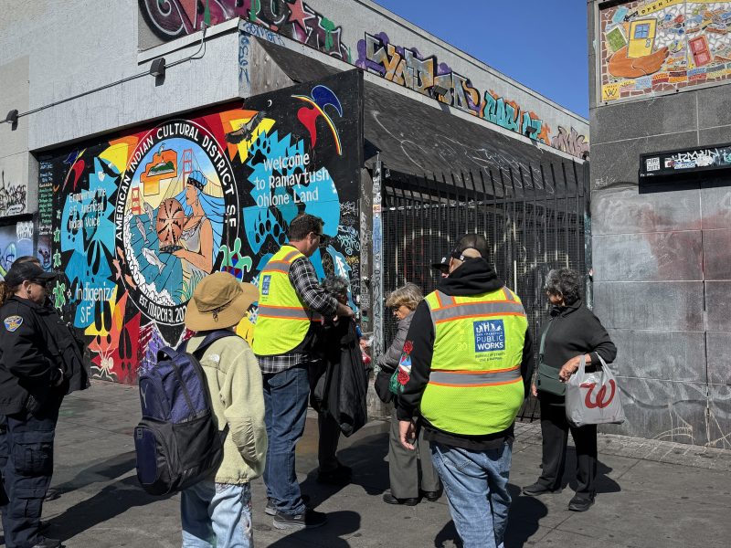 A group of people, some wearing yellow safety vests, gather near a colorful mural and graffiti-covered building in an urban area.