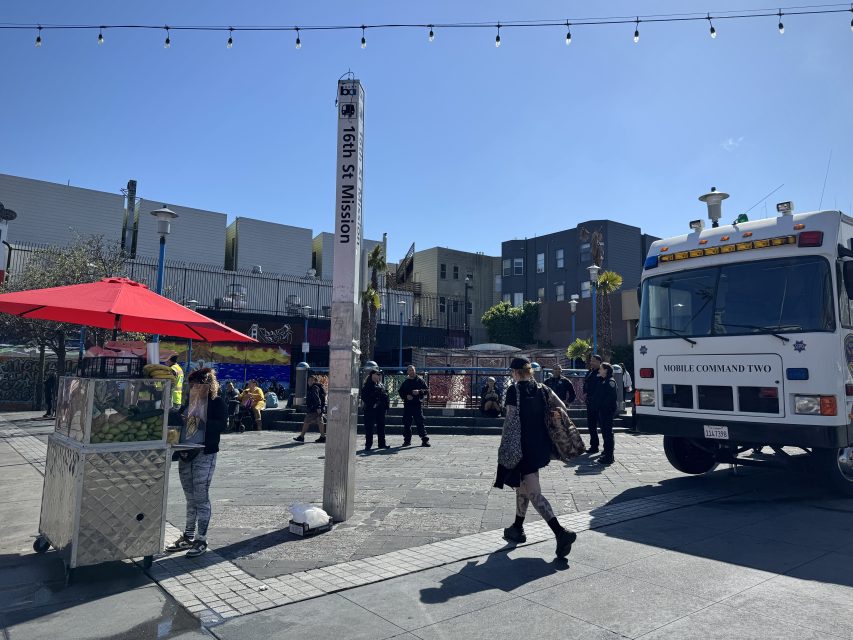 People walk through an urban plaza with a food cart, a tall street marker labeled “16th St Mission,” and a parked vehicle labeled “Mobile Command Two” on a sunny day.