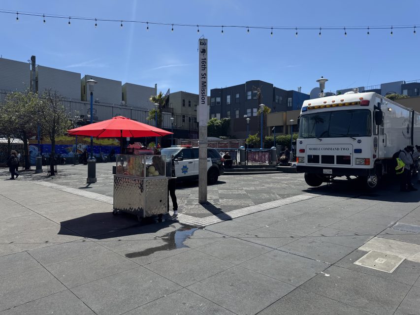 A city street scene shows a food cart with a red umbrella, a police car, a large white truck, and buildings in the background under a clear sky.
