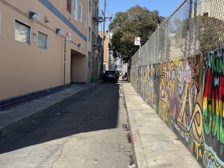 Narrow alley with parked cars and a person in the distance; left side has beige building, right side features a colorful graffiti-covered fence.
