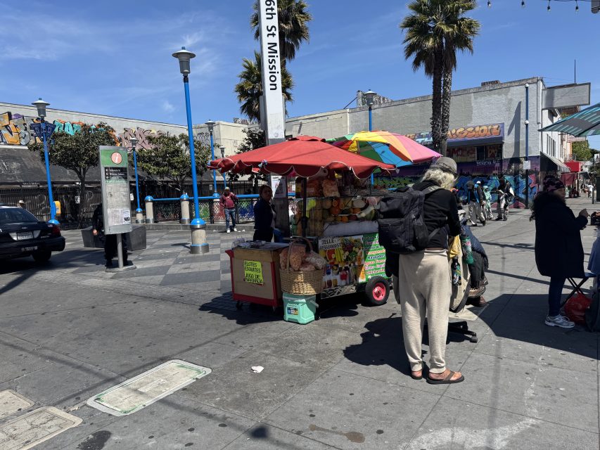 A fruit cart vendor stands under a colorful umbrella near 16th St Mission, with people walking and urban buildings in the background on a sunny day.