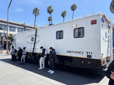 A San Francisco Police mobile command unit is parked on a city street with several people standing nearby.