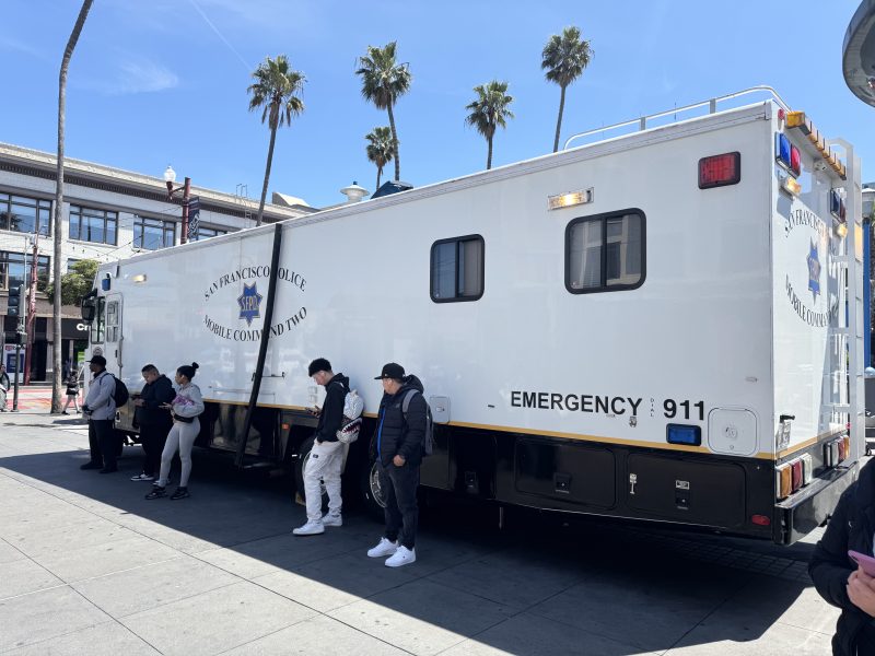 A San Francisco Police mobile command unit is parked on a city street with several people standing nearby.