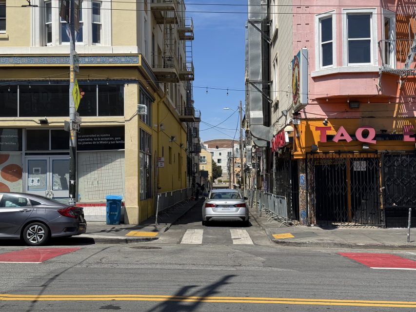 A narrow alley between two buildings with a white car parked in the middle, flanked by storefronts and a pink restaurant with partially visible signage.