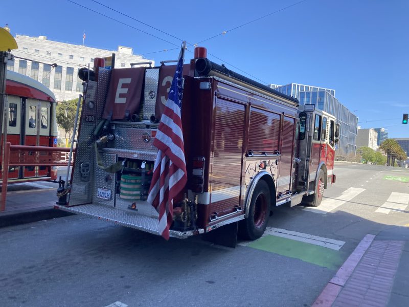 Fire truck parked on a city street near a tram, with an American flag attached to the back. Buildings and clear blue sky in the background.