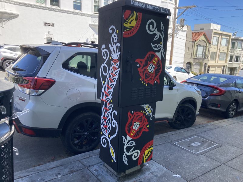Utility box on a street corner, painted with floral designs in red, white, and yellow. Cars parked nearby and residential buildings in the background.