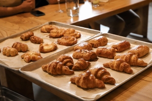 Three baking trays with a variety of golden brown croissants and pastries rest on a wooden table, with people seated in the background.