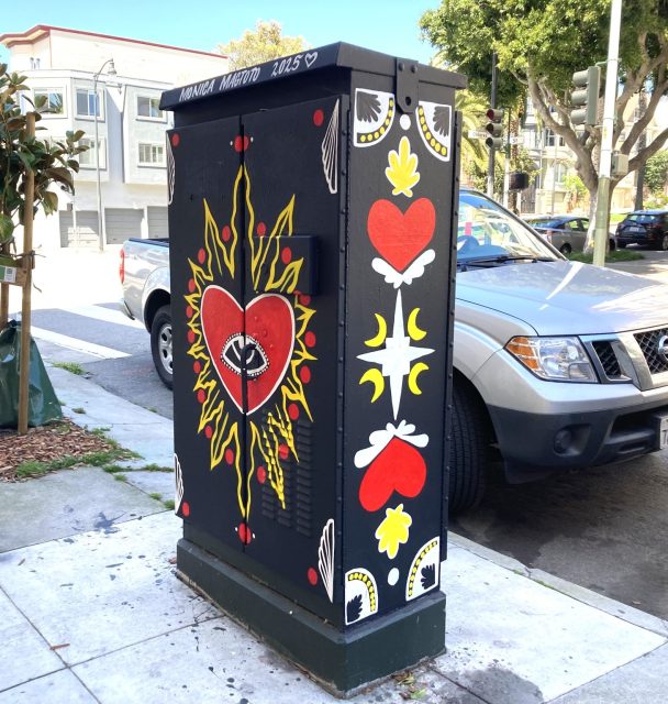 Utility box painted with red hearts, an eye inside a heart with flames, and symmetrical decorative patterns. Trees, cars, and buildings are in the background.