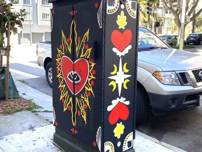 Utility box painted with vibrant designs featuring hearts, eyes, and floral motifs, labeled "Monika Mireya 2023," on a city sidewalk near parked cars and trees.