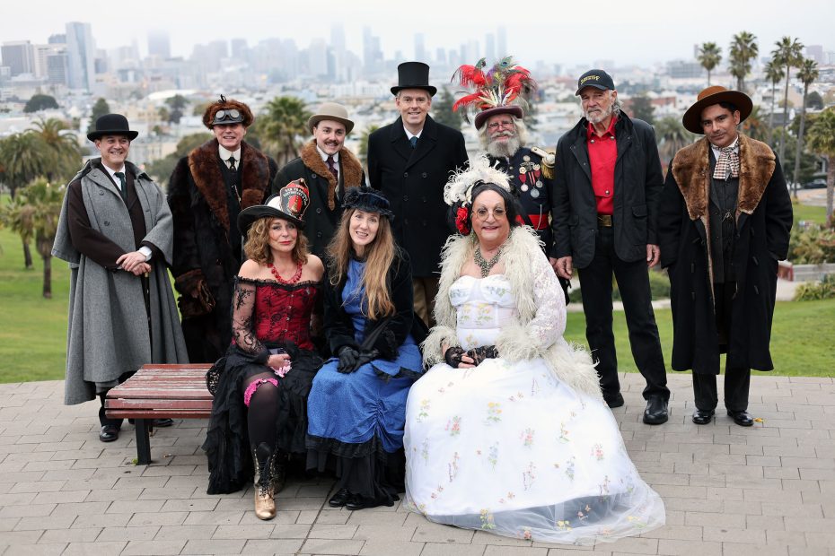 A group of people in Victorian-style costumes pose outdoors in a park, evoking the era just after the 1906 earthquake, with a city skyline in the background.