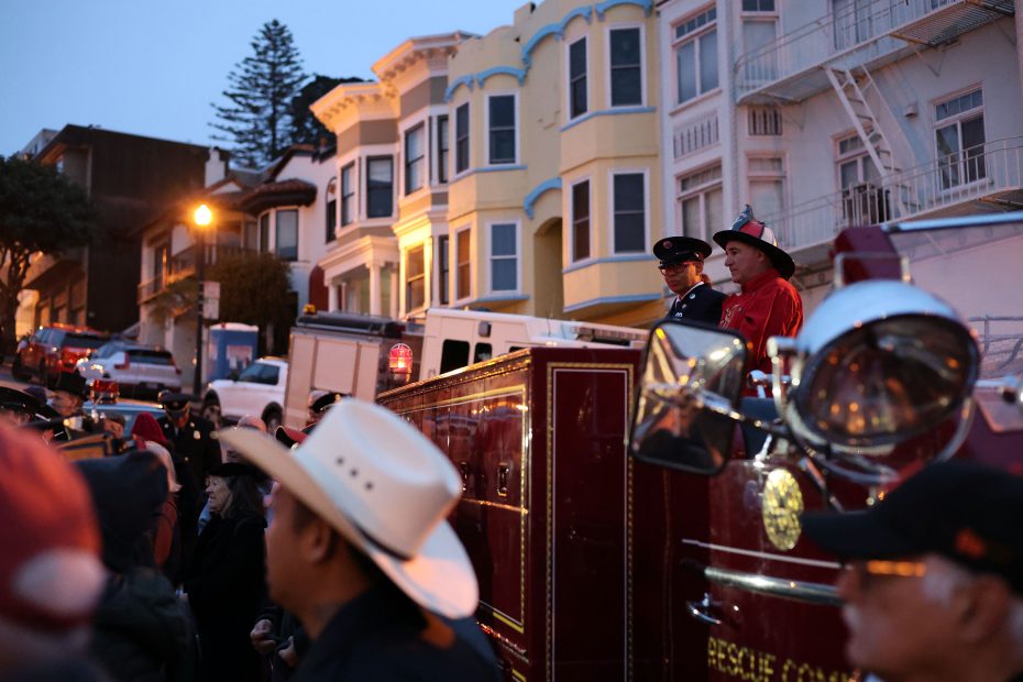 A crowd gathers near vintage fire trucks with uniformed personnel in front of colorful, multi-story residential buildings at dusk, evoking scenes reminiscent of the 1906 earthquake response.