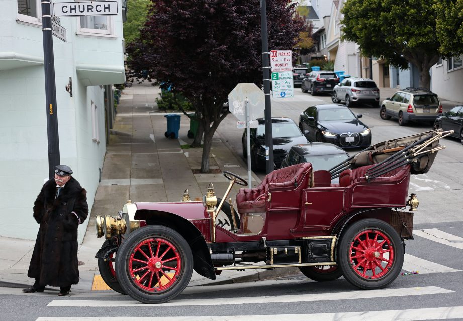 A person in a fur coat and hat stands on a street corner near a vintage red car with brass details and red wheels, evoking the era just after the 1906 earthquake, parked at an intersection.