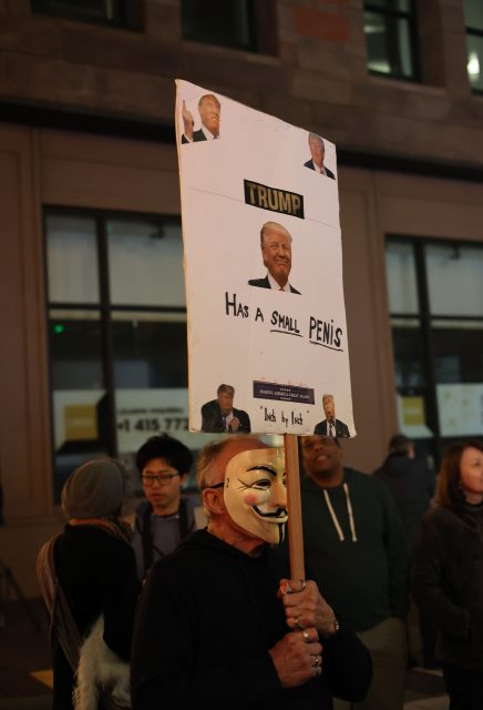 A person wearing a Guy Fawkes mask holds a protest sign with images of Donald Trump and text making a derogatory statement about him, drawing parallels to the chaos of the 1906 earthquake. Other people stand nearby on a city street at night.