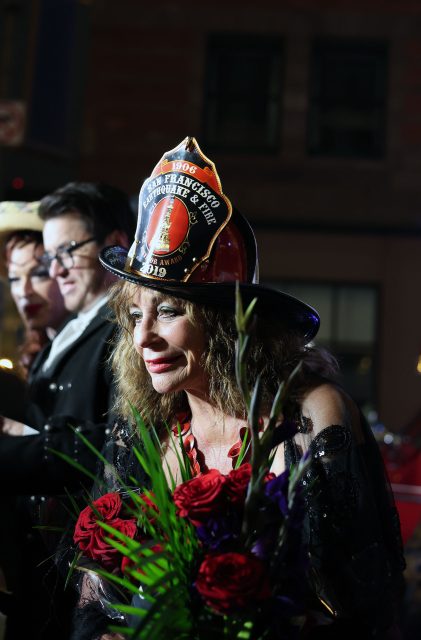 A woman in a black outfit and a firefighter hat holds a bouquet of red roses, standing outdoors at night among others, evoking the spirit of bravery seen during the 1906 earthquake.